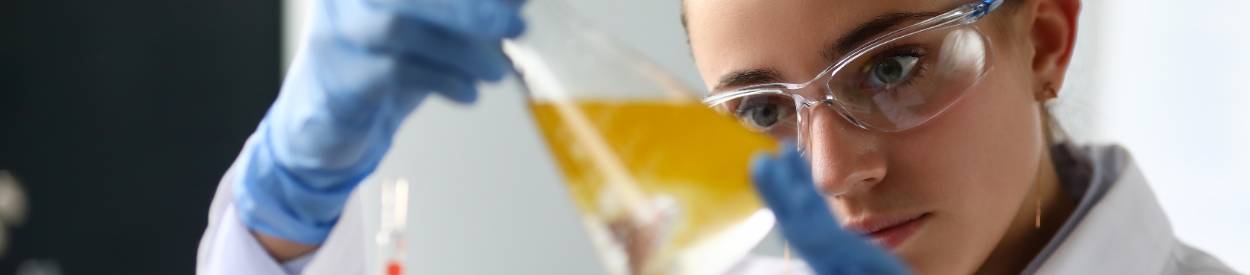 Close-up of serious laboratory worker holding ampoule in front of eyes and examines contents. Scientist in protection gloves eyewear and white coat.